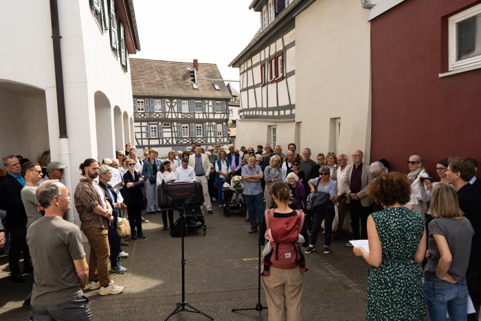 Frau redet am Mikrofon. Menschengruppe hört zu